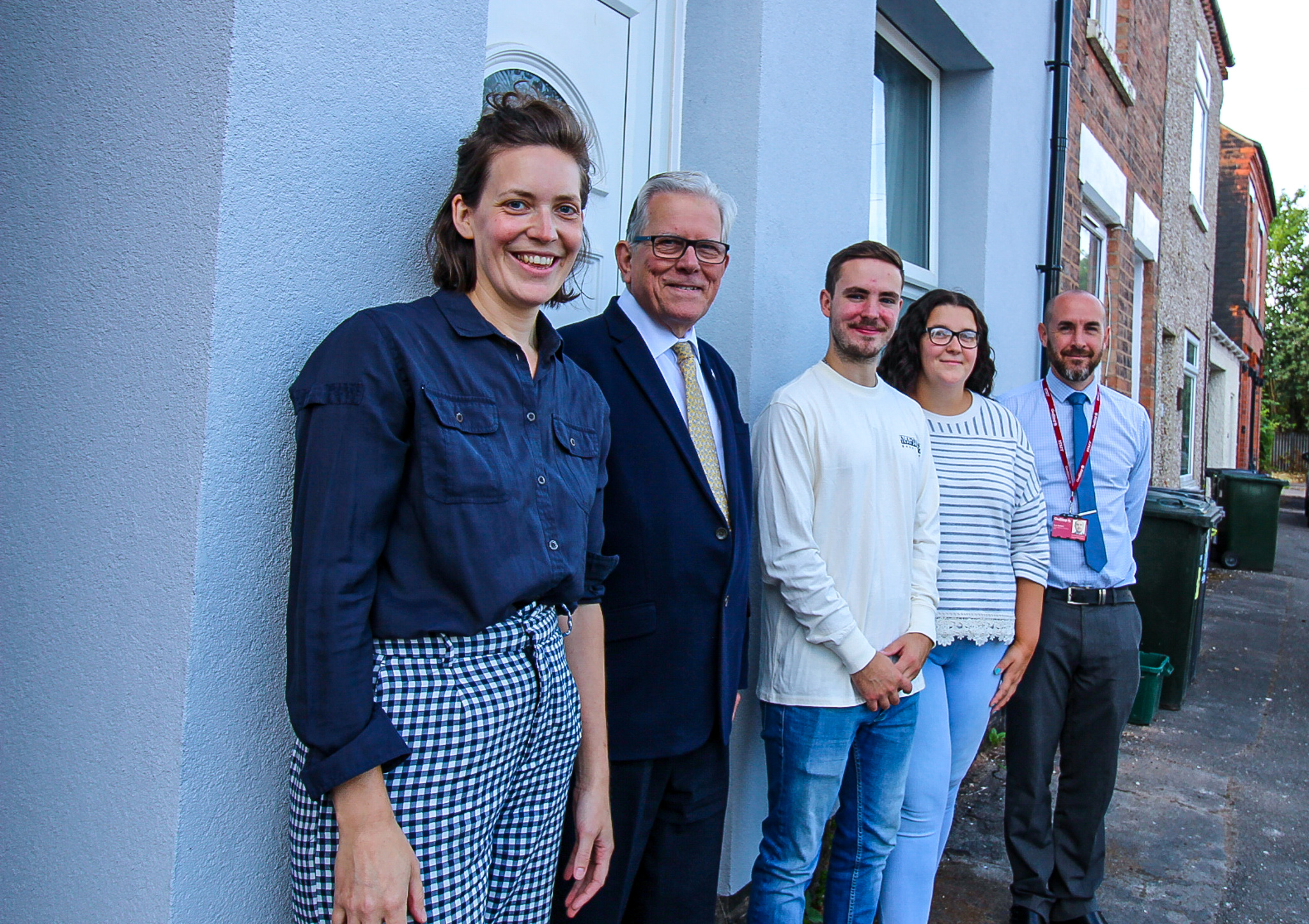 A group of people stood in front of a newly rendered house. The outside of the house is blue/purple in colour and has had external wall insulation installed.